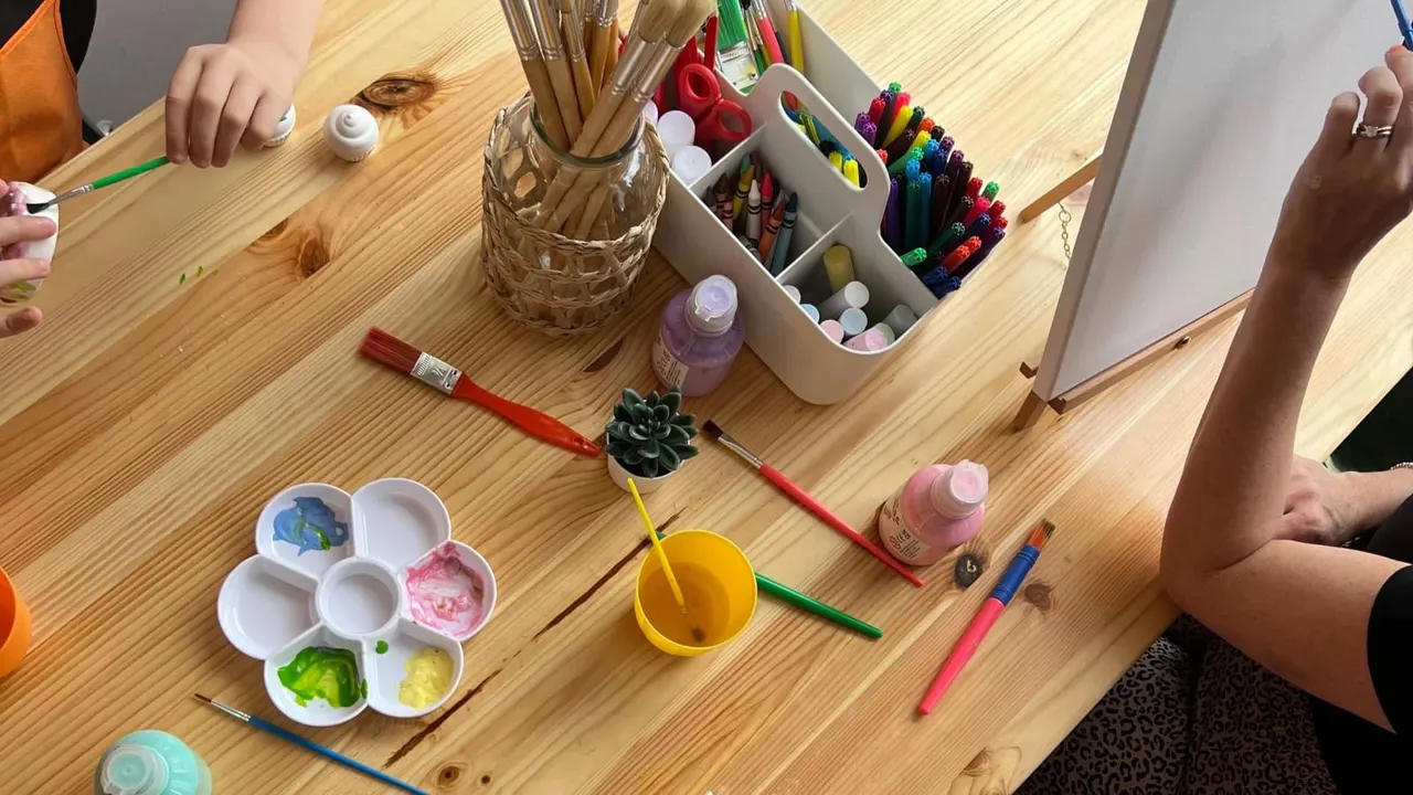 Children engaged in an arts and crafts activity at a wooden table with paints, brushes, markers and creative supplies.