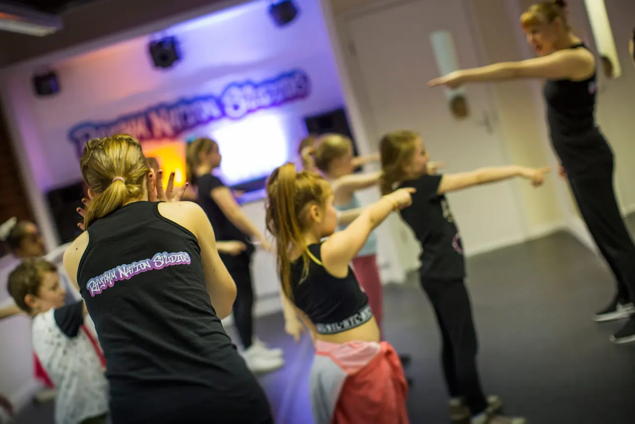 Children in black dance attire follow an instructor's movements during a dance class in a studio with purple lighting.