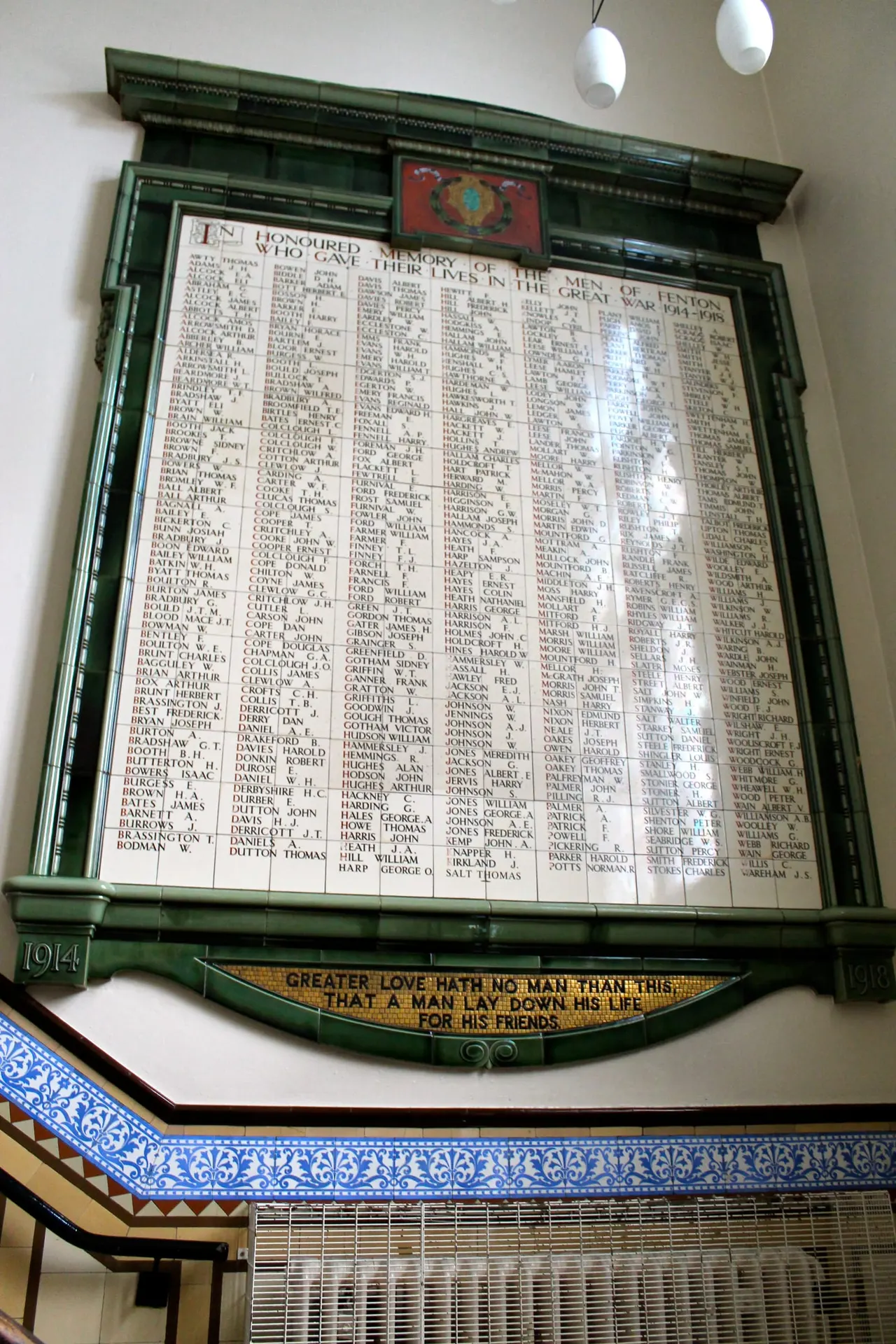 A First World War memorial mounted on a white wall listing names of men who died, with a green wooden frame and a biblical inscription below.