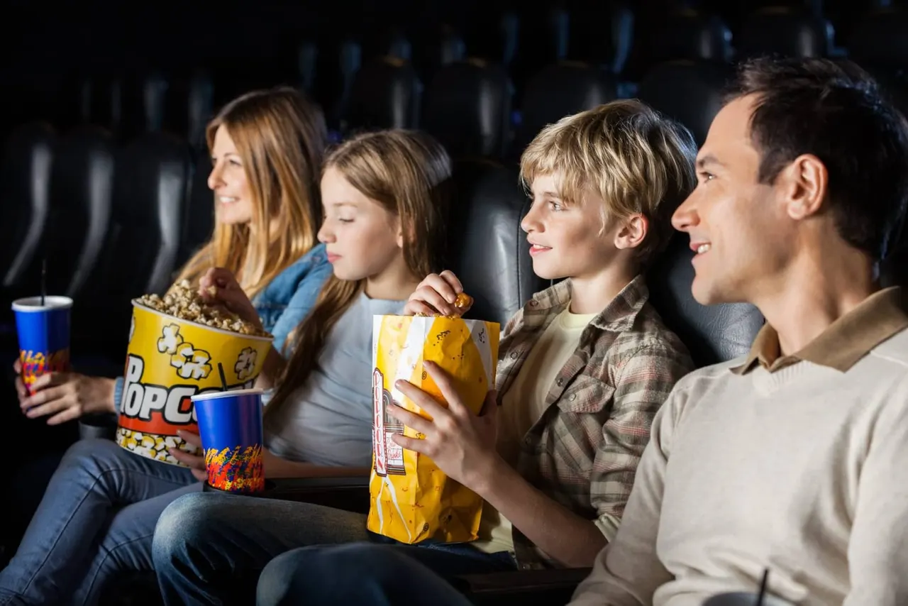 Family of four watching a film in a cinema, holding popcorn and drinks.