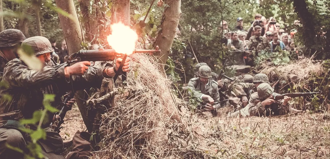 Costumed participants in military uniforms firing weapons with spectators watching from the sidelines in a wooded area.