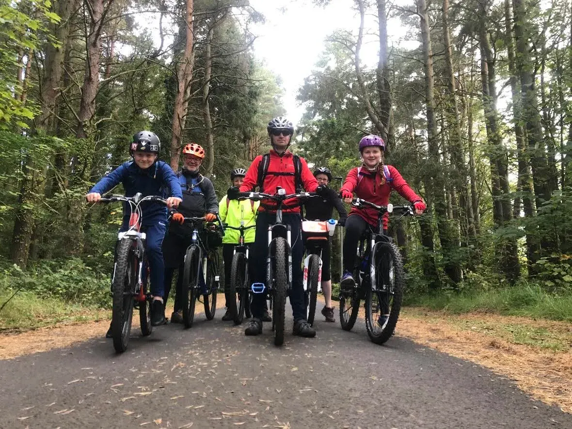 A group of five cyclists wearing helmets riding on a forest trail lined with tall trees.