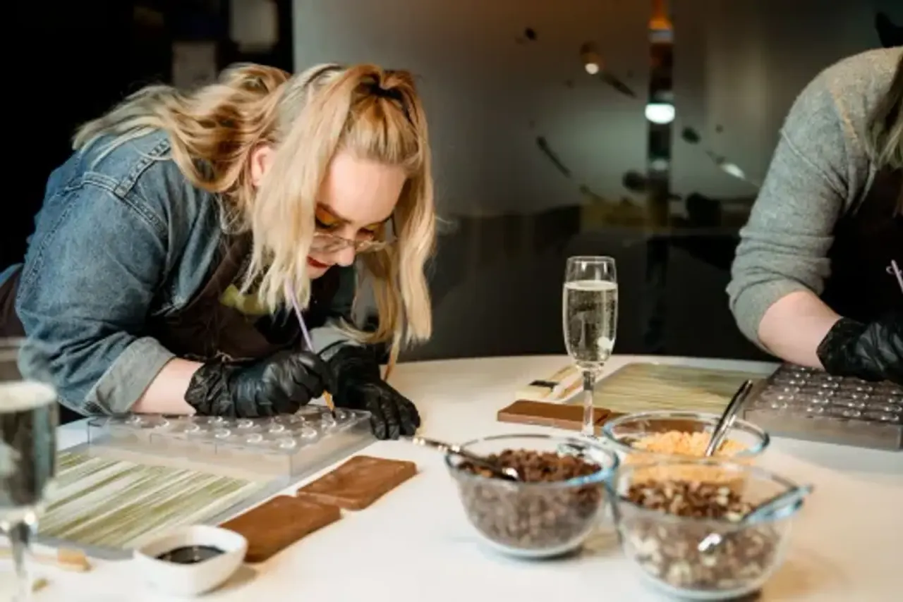 Woman in denim jacket and black gloves examining chocolate samples at a tasting table with bowls of ingredients.