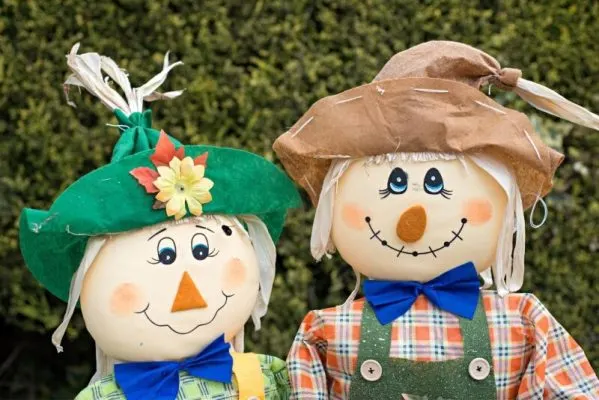 Two painted scarecrow figures with smiling faces, wearing hats and blue bows, positioned against a green hedge backdrop.