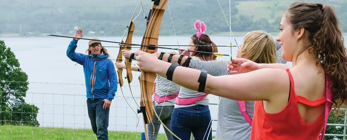 Young people practicing archery outdoors with an instructor, lake and hills visible in the background.