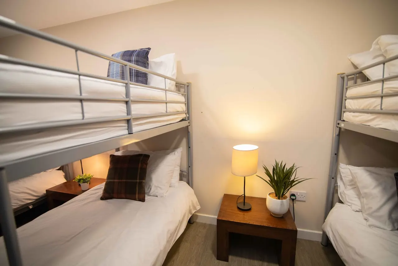 A compact dormitory room with metal-framed bunk beds on either side, white bedding, a central wooden table with a lamp and potted plant, and warm overhead lighting.