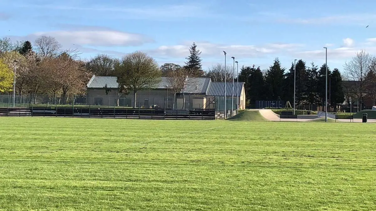 Empty sports field with pavilion buildings, fencing and evergreen trees under blue sky.