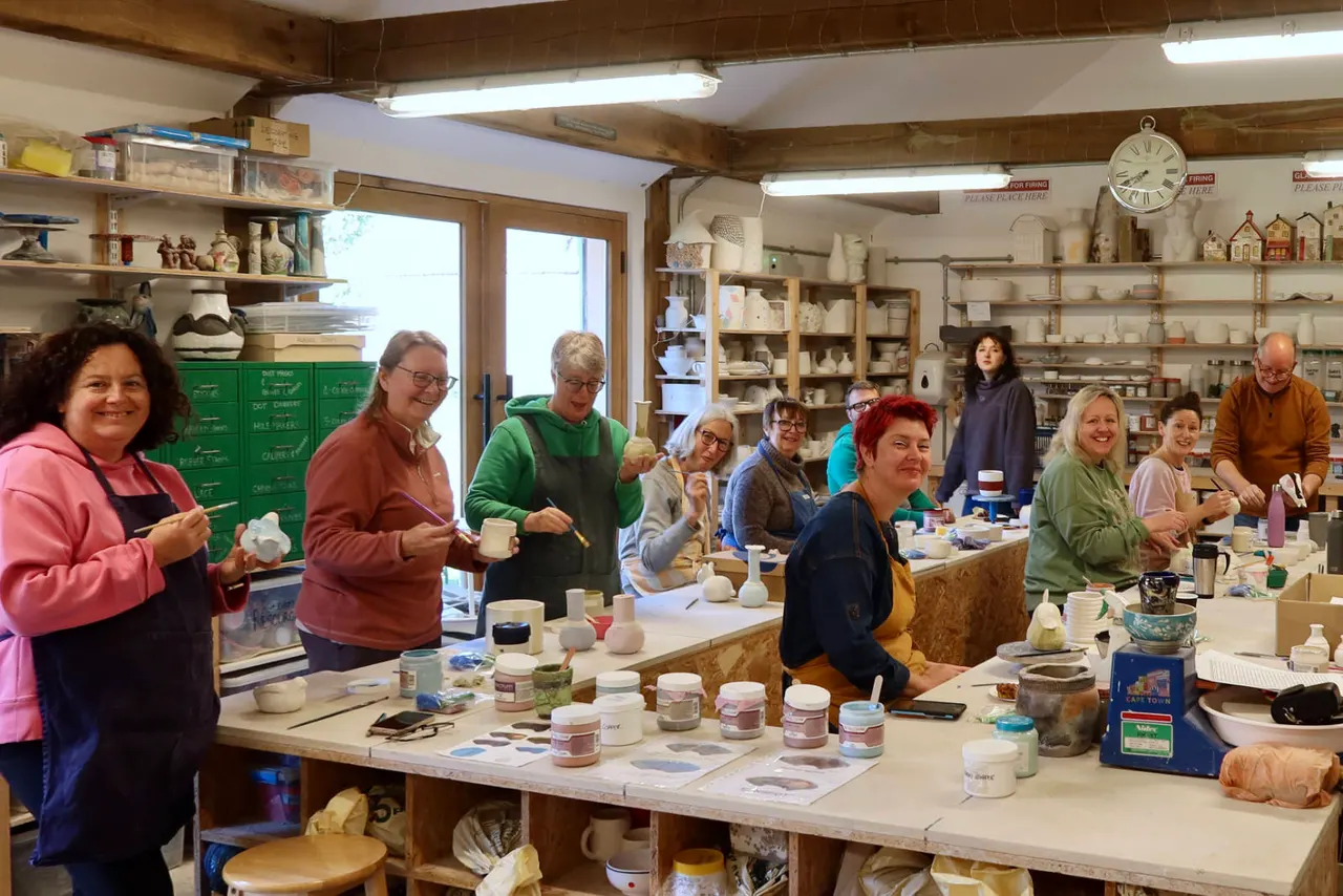 A group of adults work on pottery projects together at tables in a studio workshop surrounded by shelves of ceramic supplies and finished pieces.
