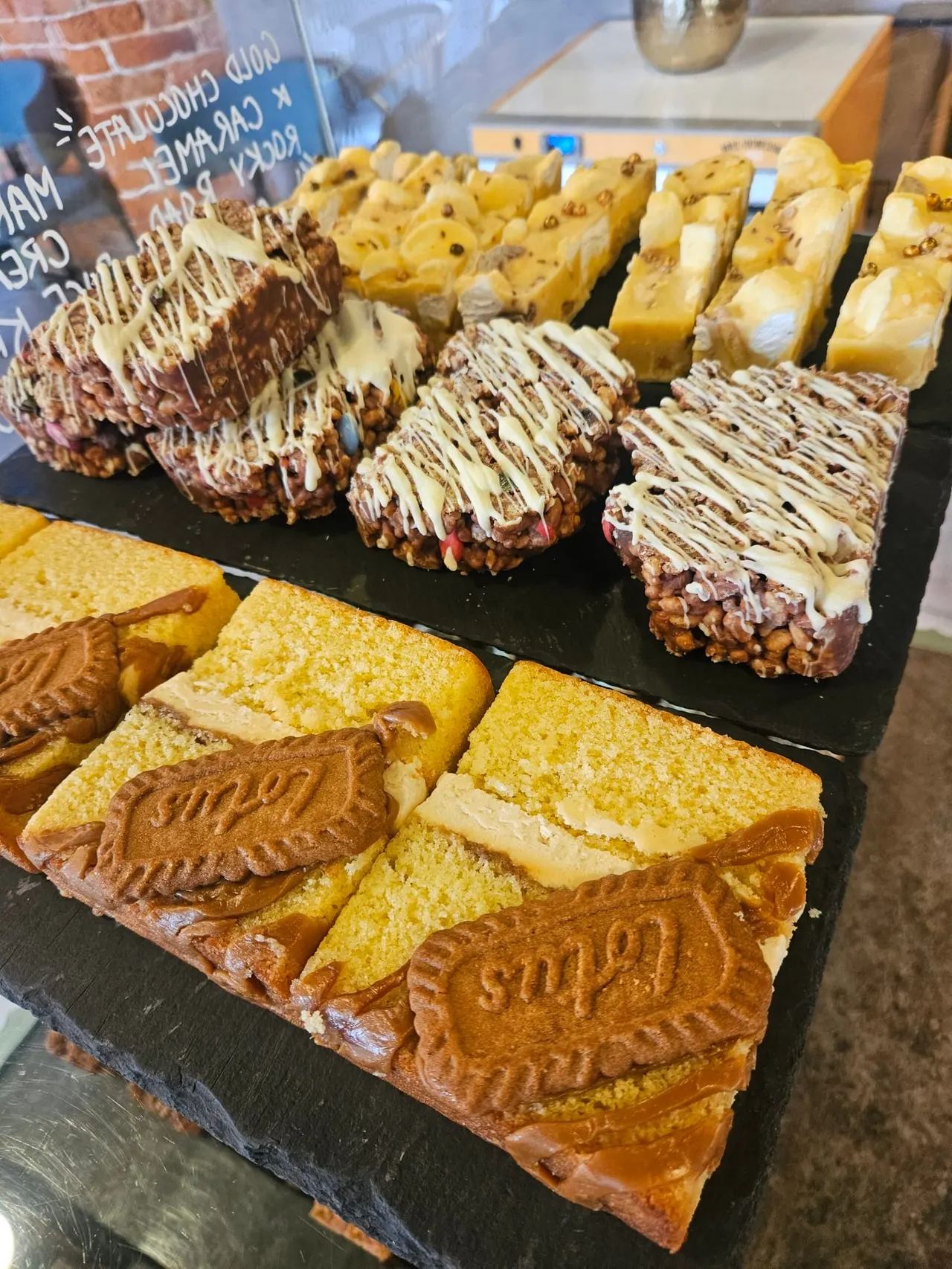 A display of homemade cakes and bakes including chocolate drizzled brownies, flapjacks, and frangipane tarts on a black slate board.