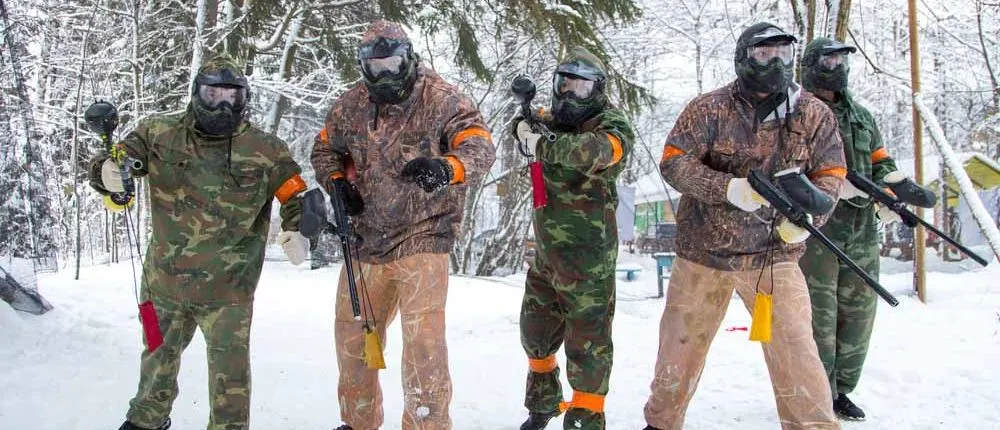 Six paintball players in protective gear and masks stand together in snowy woodland during a game.