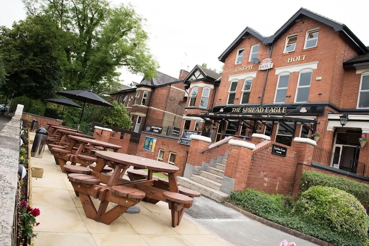 Pub garden with wooden picnic tables on a paved terrace, brick buildings and green trees in background.