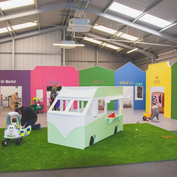 Young children explore a brightly coloured indoor play area with a mock campervan, toy vehicles, and themed activity zones.