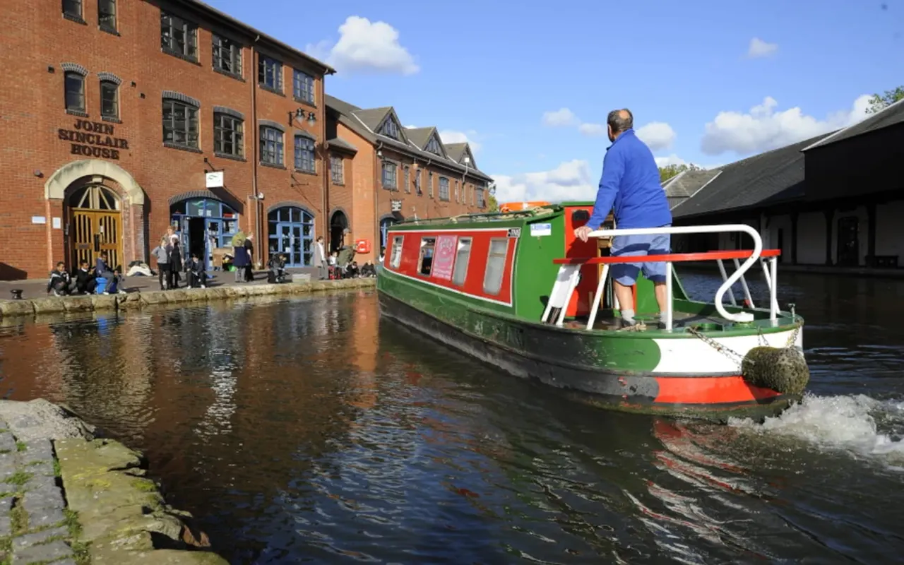 Adult and child on a colourful narrowboat moored at a canal basin beside a brick warehouse building.