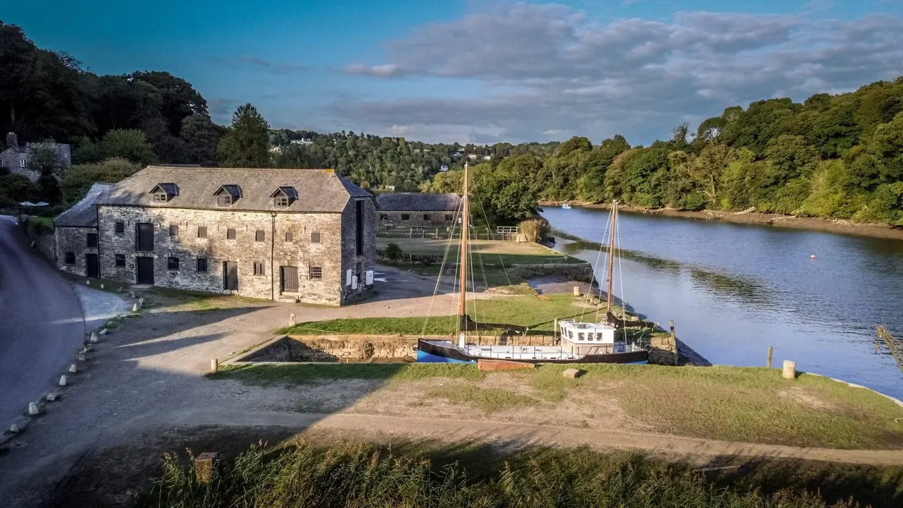 Stone riverside buildings with moored sailing boat on tidal estuary surrounded by woodland.