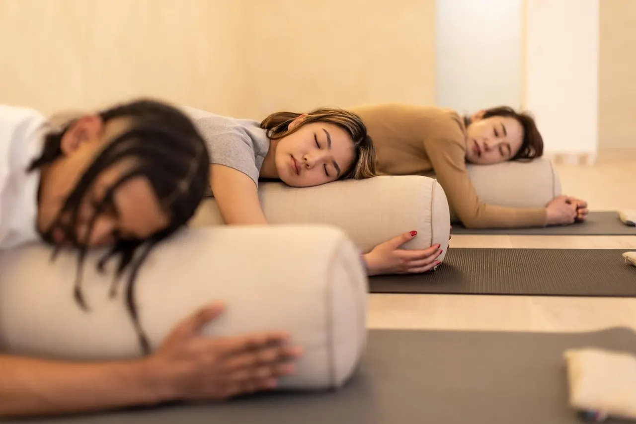 Three people lying on yoga mats with bolsters during a guided relaxation or restorative yoga class.