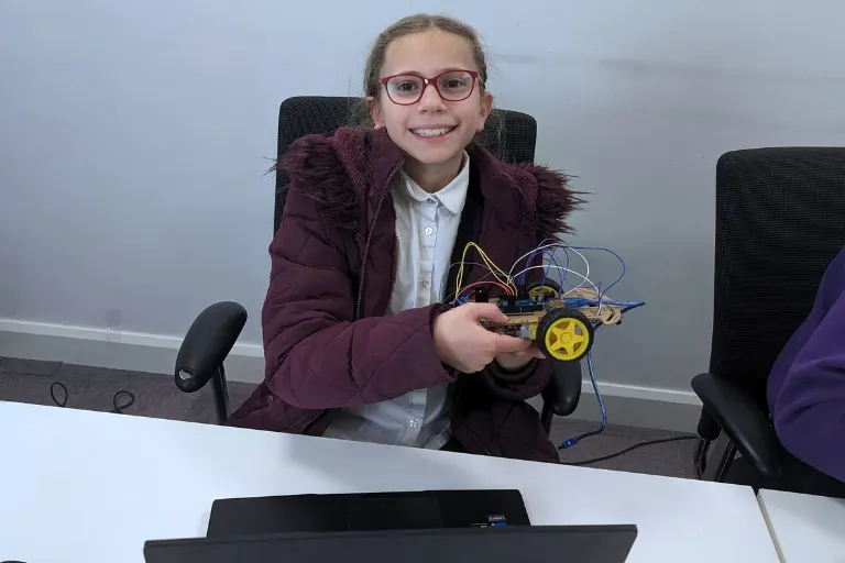 Girl holding a small robot vehicle at a desk during a coding or robotics activity.