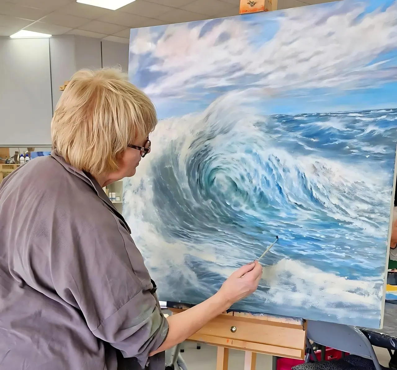 Woman painting a seascape with crashing waves on canvas in an indoor studio setting.