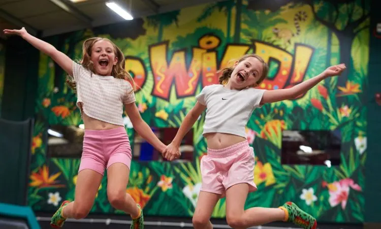 Two young girls jumping on a trampoline with arms outstretched against a colourful jungle-themed wall.