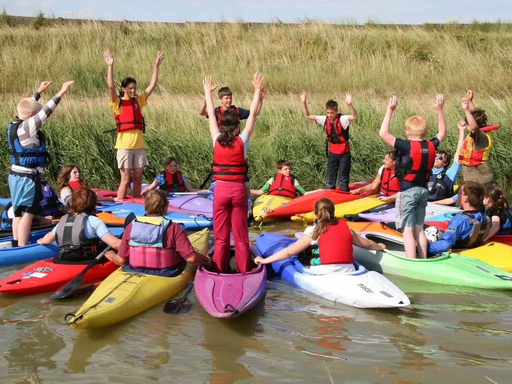 Group of children in life jackets gathered around colourful kayaks in shallow water, arms raised, with reed beds behind.