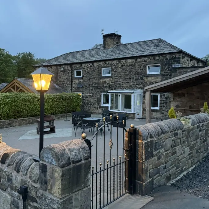 Stone farmhouse with illuminated lamp post and seating area on paved terrace at dusk.