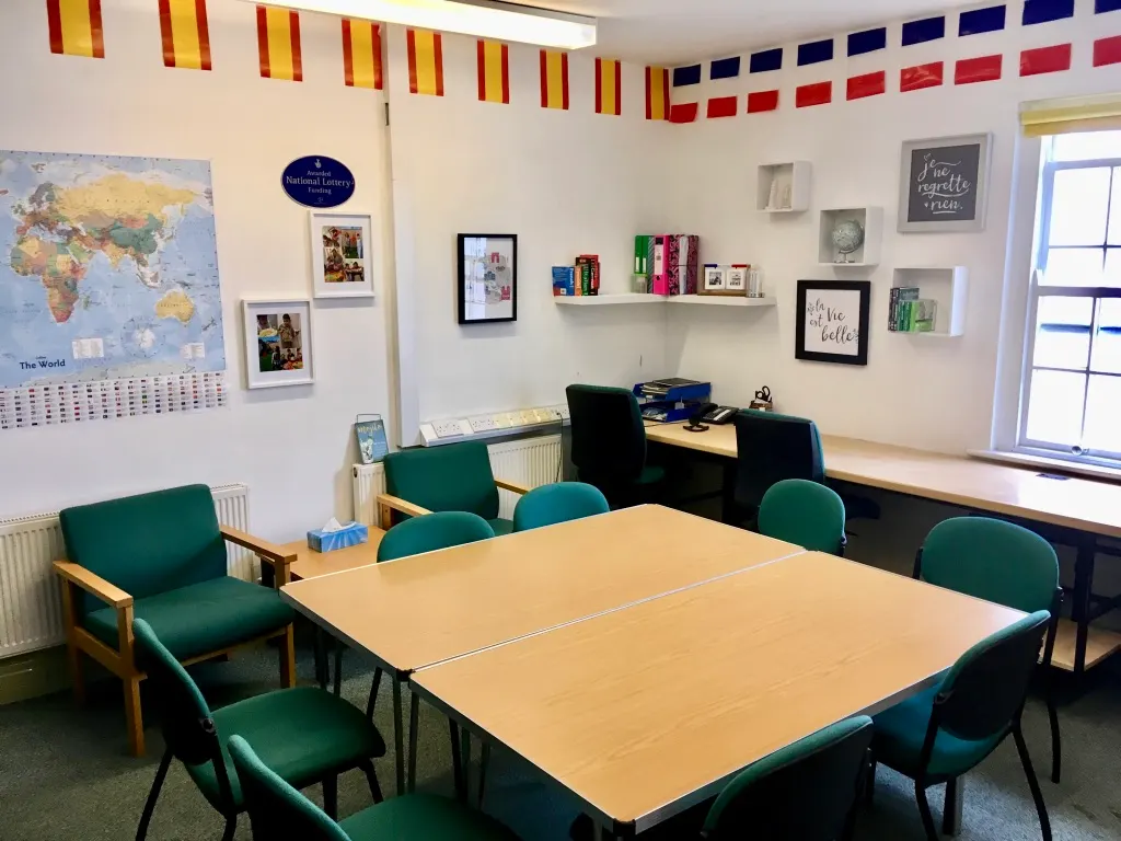 A classroom interior with a large wooden table, green and black chairs, world map, shelving with books, and international flags on the walls.