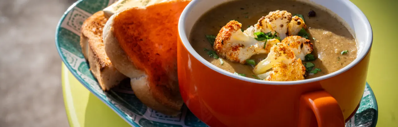 A bowl of creamy soup with roasted cauliflower florets and toasted bread on the side, served in a red bowl.