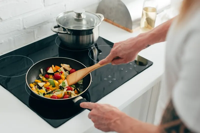 Person stirring coloured vegetables in a frying pan on an induction hob in a modern kitchen.