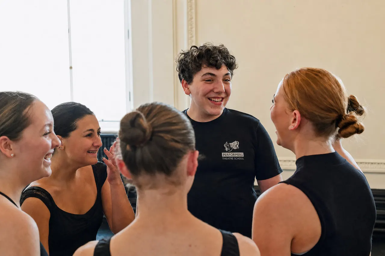 A group of young people in black dance wear laugh together during a theatre school class or rehearsal.