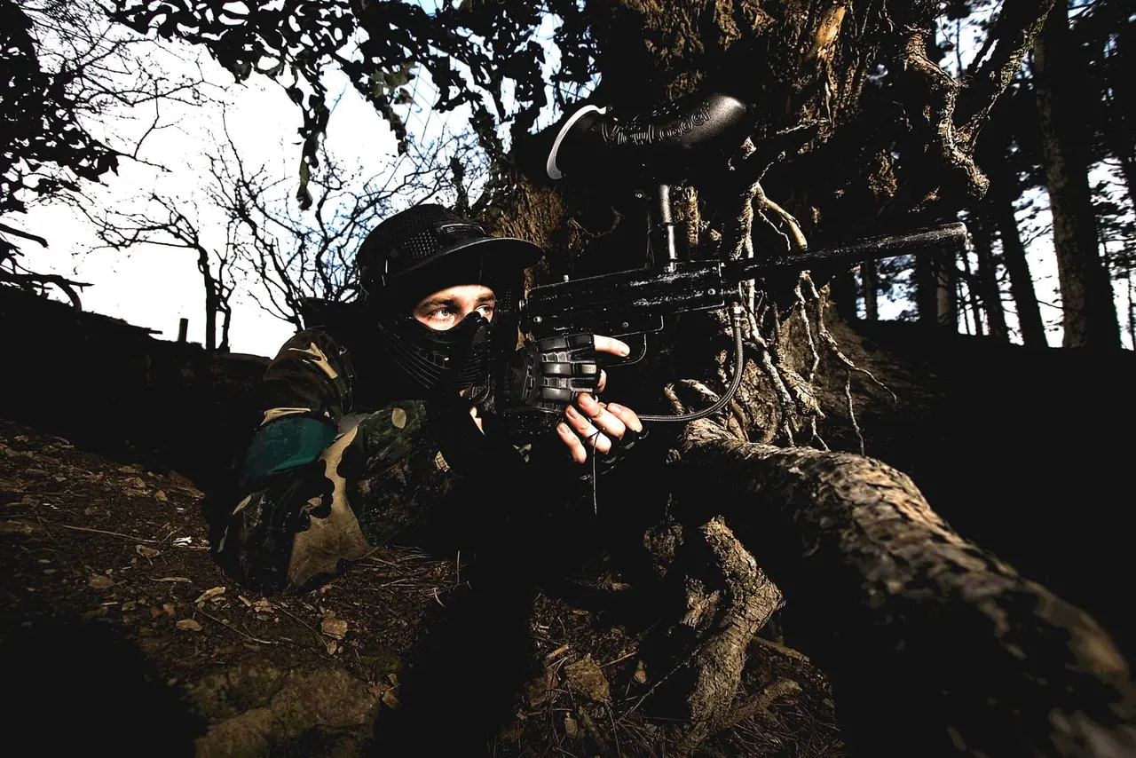 Player in paintball gear holding marker gun in woodland setting amongst trees and logs.