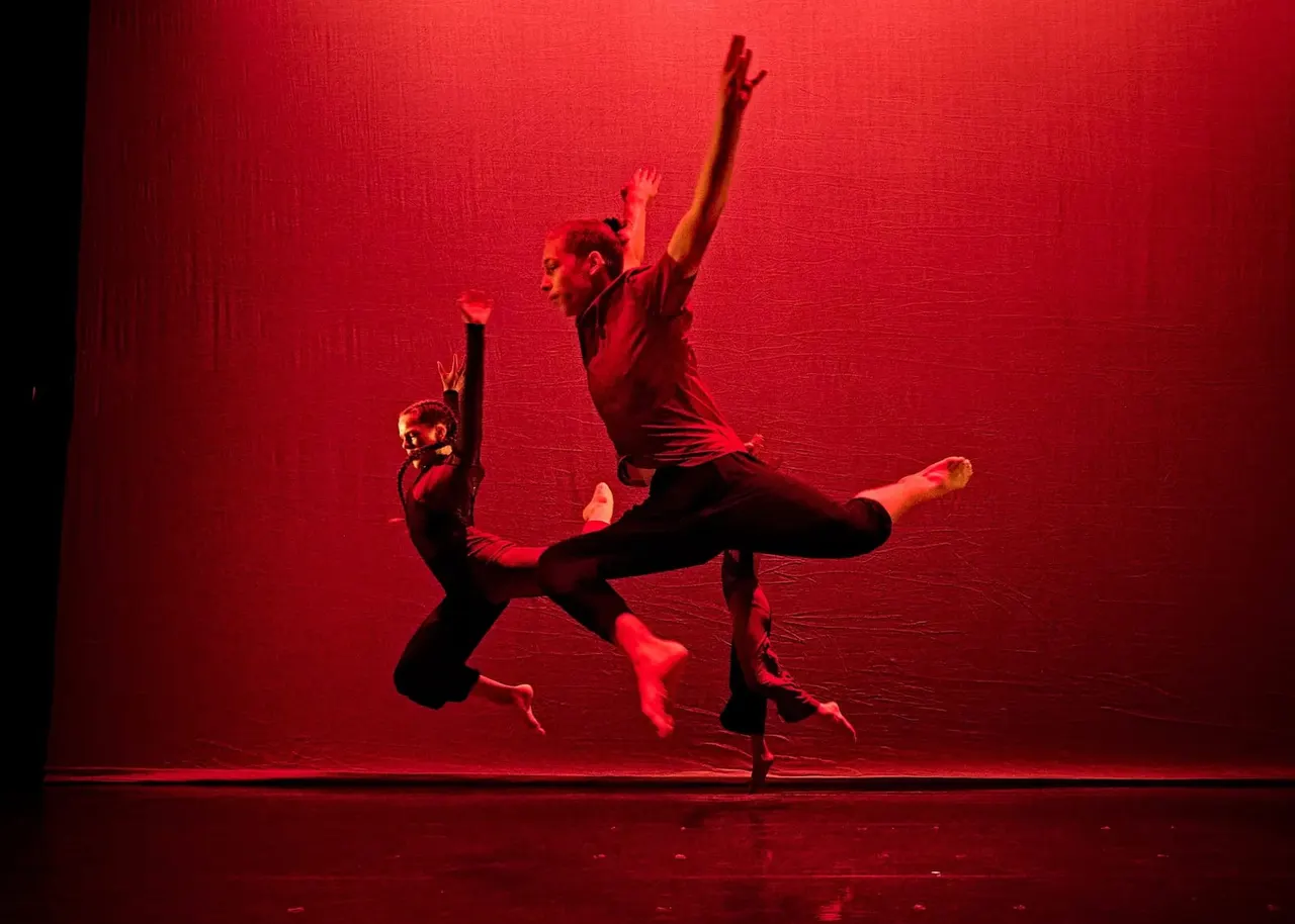 Three dancers performing a contemporary dance piece on stage with dramatic red lighting and backdrop.