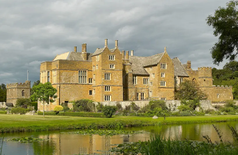 Stone castle with multiple turrets and chimneys beside a moat under cloudy sky, surrounded by green lawns and trees.