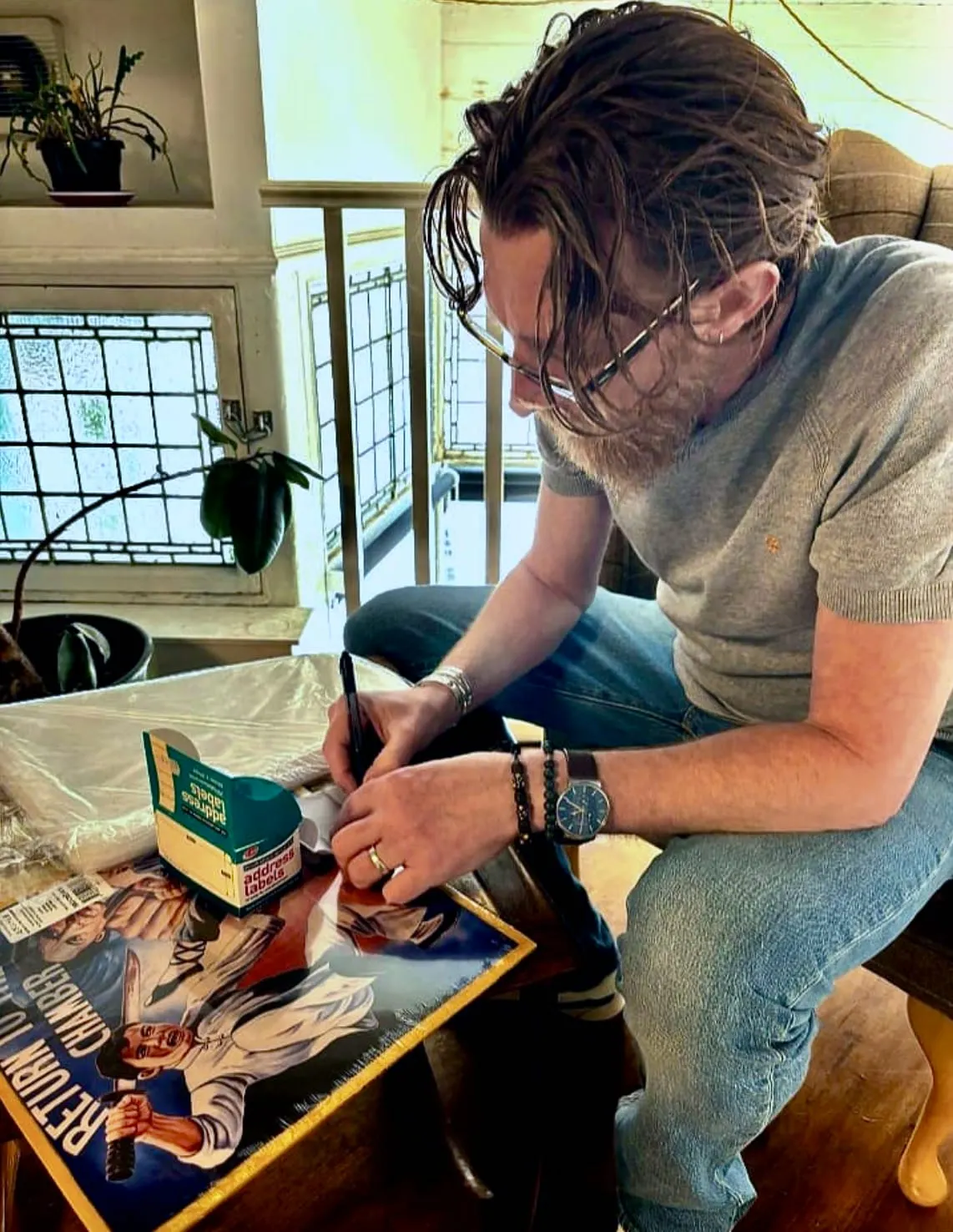 Man signing a poster indoors beside potted plants and windows.