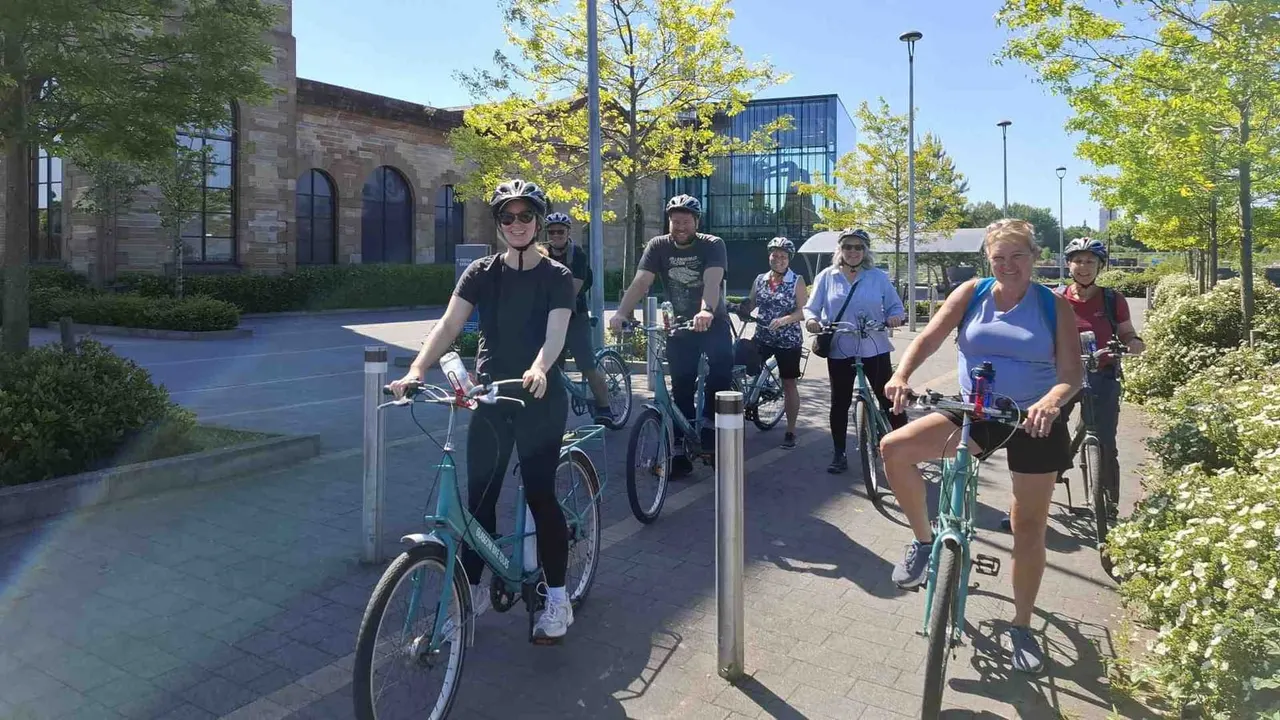 A group of cyclists wearing helmets ride along a paved path past modern buildings and green trees on a sunny day.