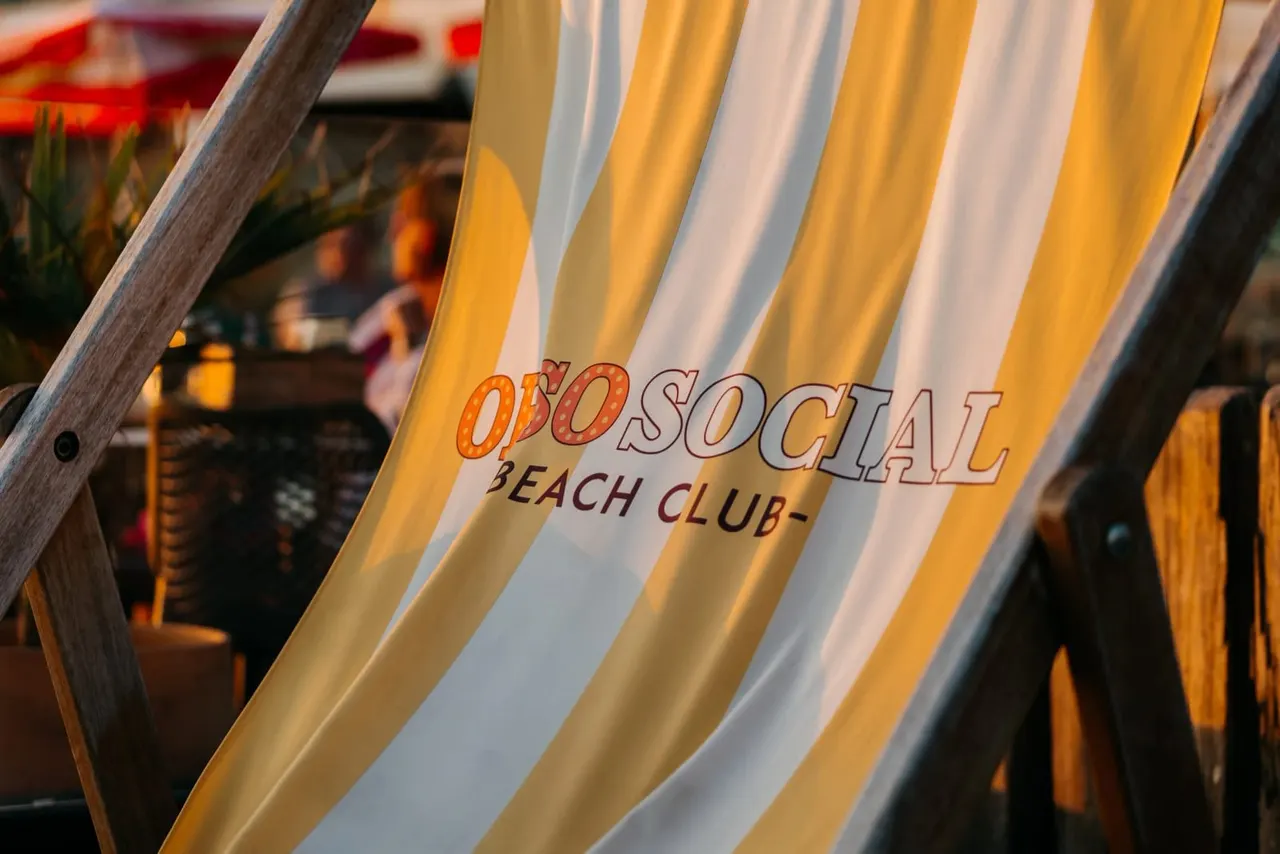 Close-up of yellow and white striped deckchair with "OHSO Social Beach Club" text, with blurred beach venue and people in background.