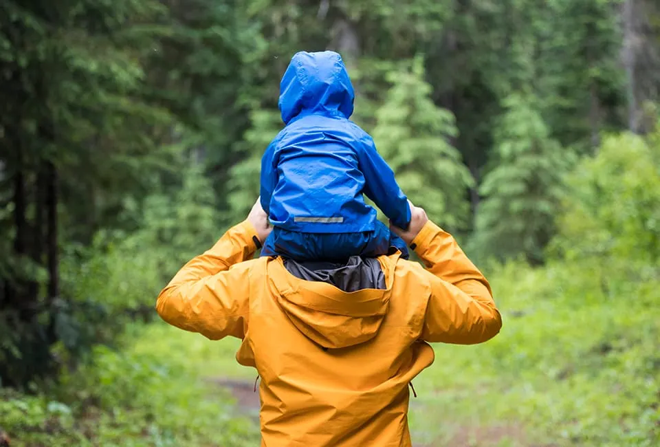 Child in yellow jacket and blue backpack walking on a woodland path surrounded by green trees.