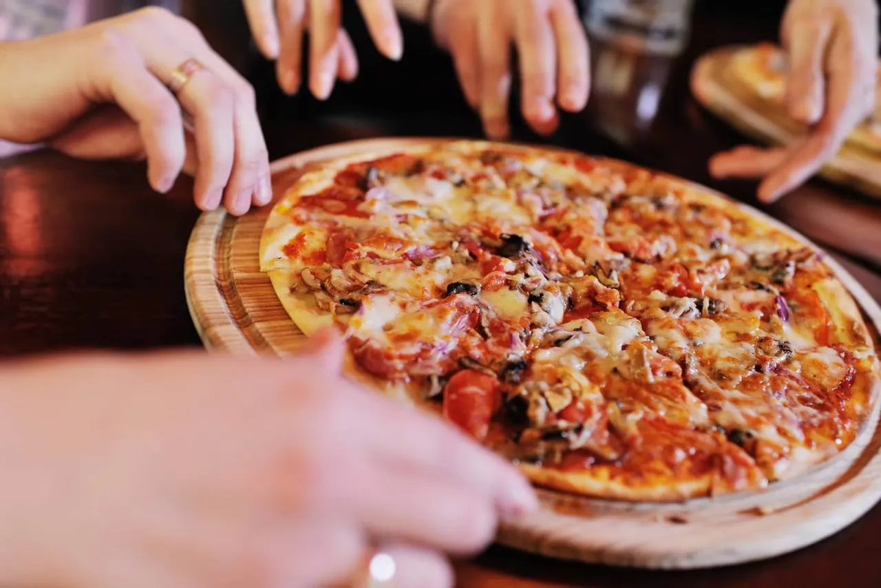 Multiple people reaching towards a freshly baked pizza on a wooden board.