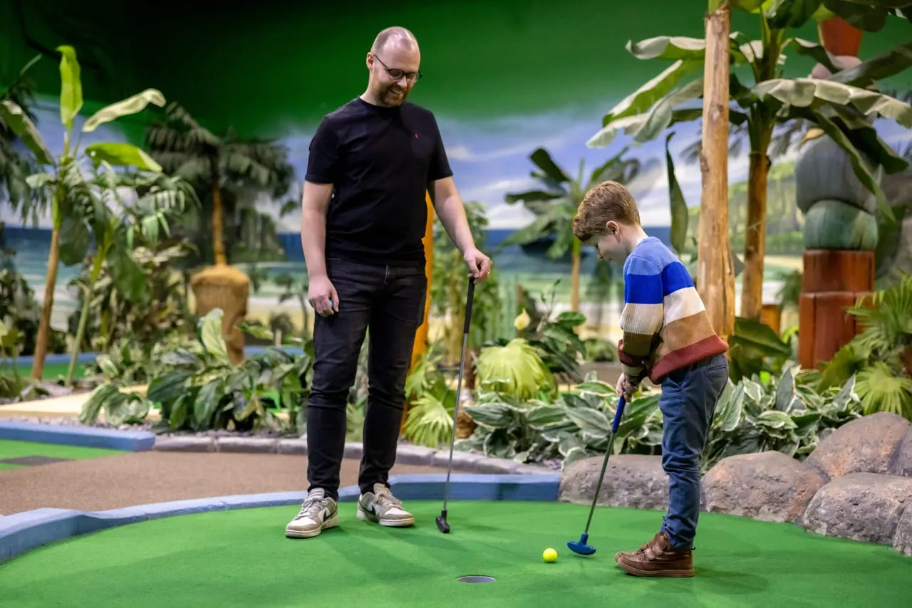 Adult and child playing mini golf surrounded by tropical plants and themed scenery.