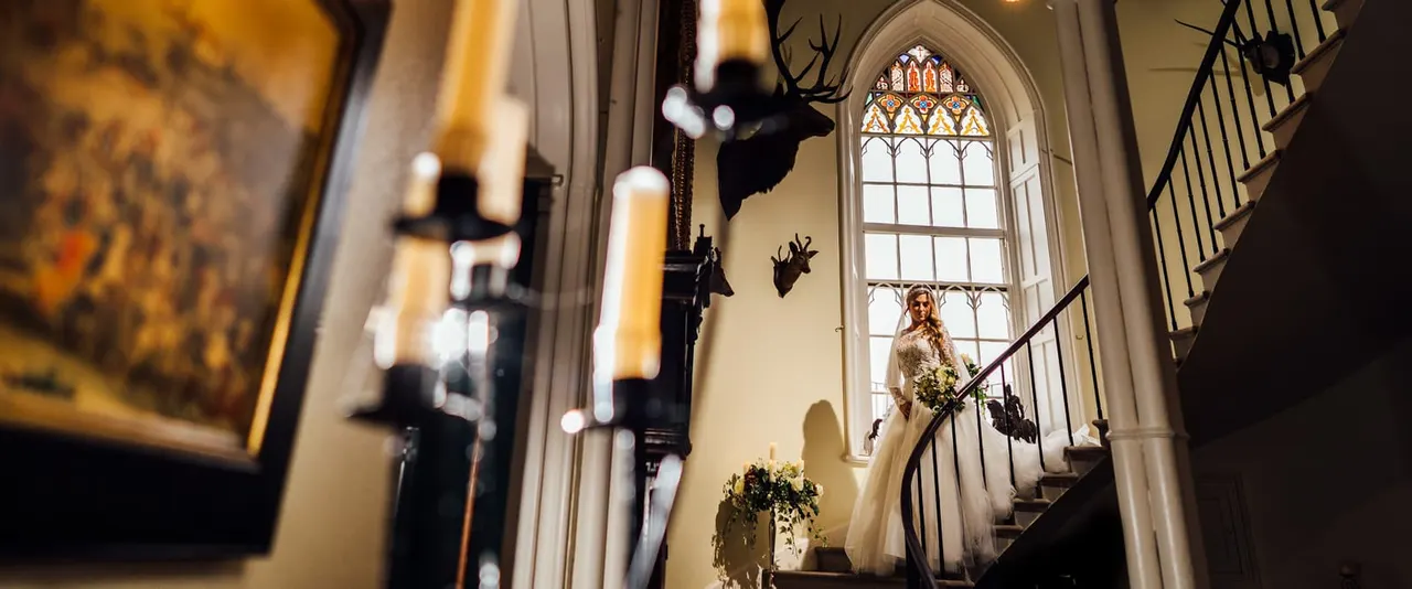 Bride descending ornate staircase in grand hall with stained-glass window and decorative animal heads on walls.