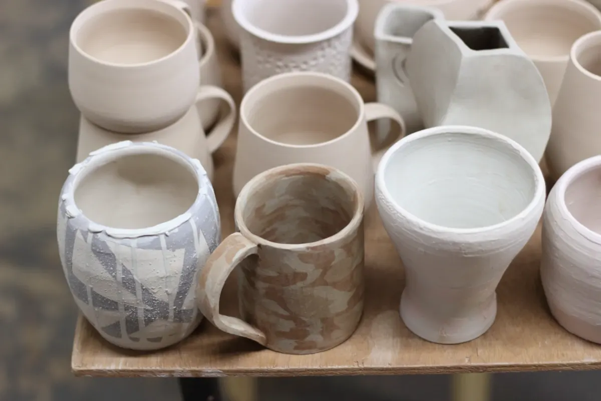 Collection of handmade ceramic cups, mugs and jugs in white and neutral tones displayed on a wooden shelf.