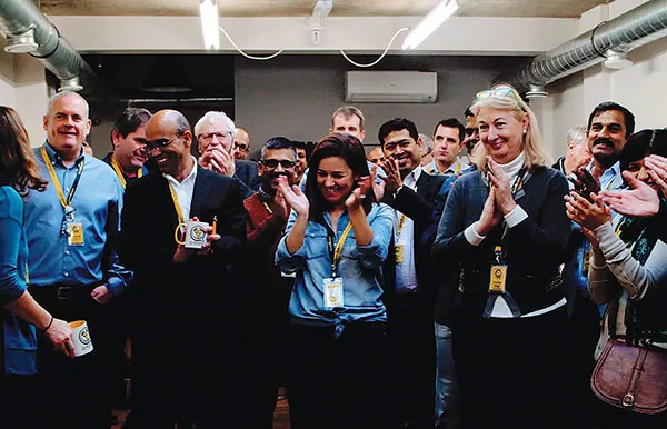 A group of diverse people wearing lanyards and ID badges stand together clapping and smiling in an indoor venue.