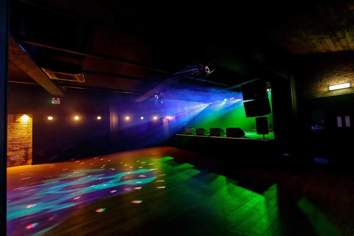 Empty nightclub interior with colourful stage lighting in blue, green and purple, dance floor, and DJ booth.