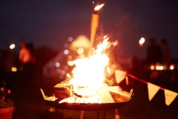 People gathered around a blazing bonfire at night with bunting visible nearby.