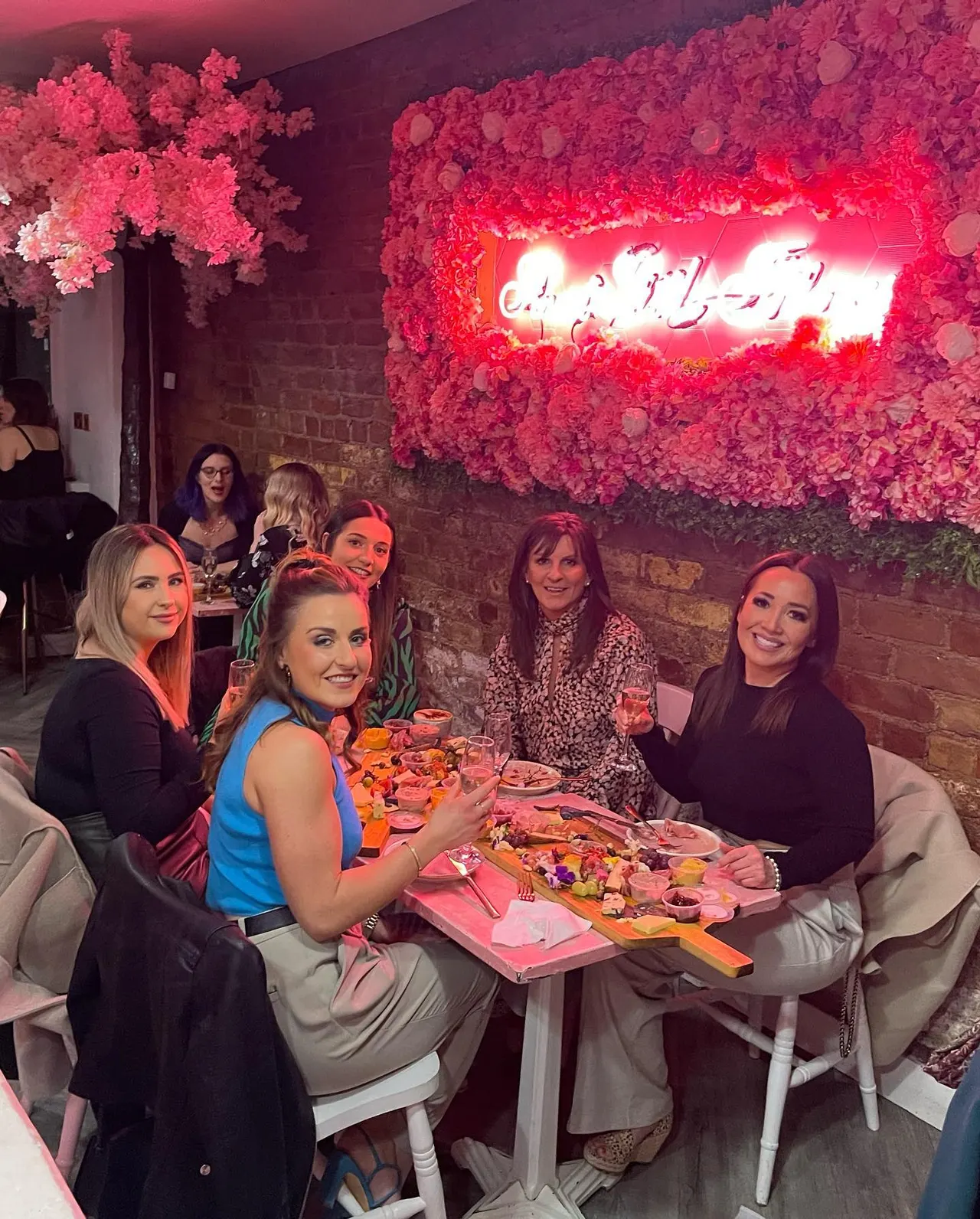 Group of women dining at a table with food and drinks under pink neon sign and floral decorations.