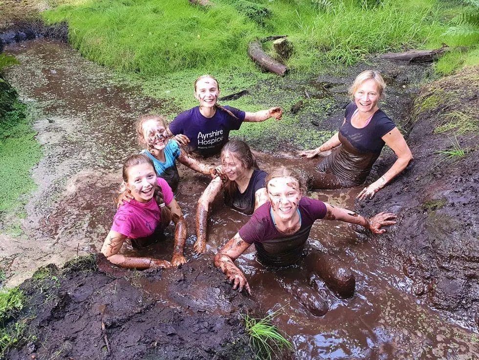 Adults and children playing and covered in mud in a shallow stream surrounded by grass and moss-covered rocks.