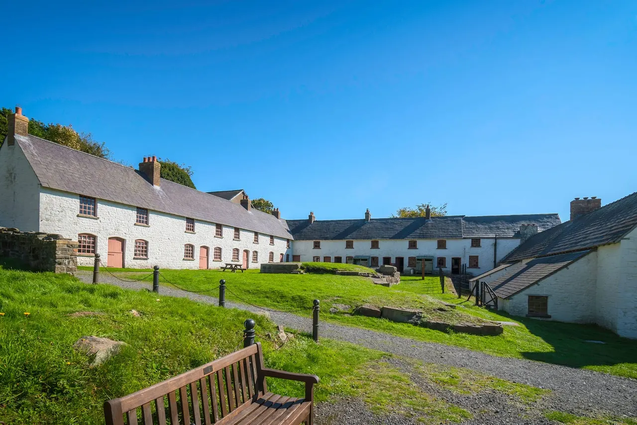 Historic whitewashed stone buildings arranged around a grassed courtyard on a clear sunny day.