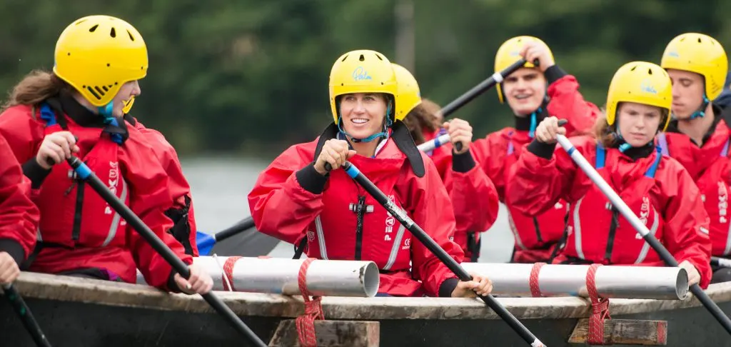 Group of young people in red jackets and yellow helmets paddling a raft on water.