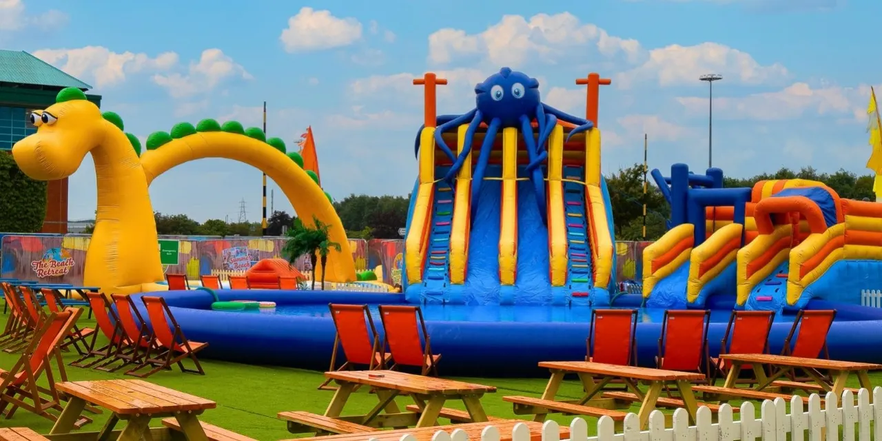 Inflatable bouncy castle attractions with bright blue and yellow slides in a leisure park, surrounded by deckchairs on grass.
