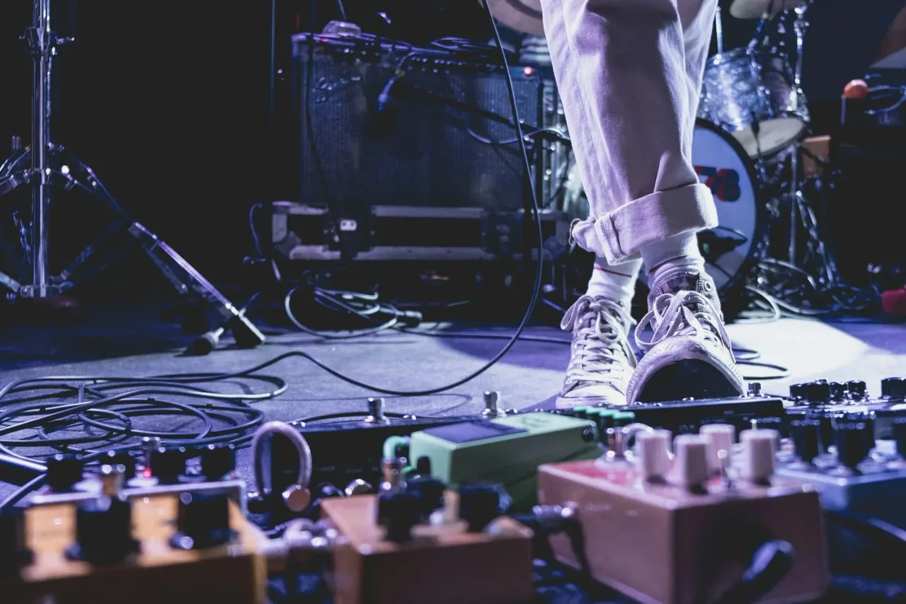 Musician performing on stage wearing white trainers, standing at audio equipment and microphone with band setup visible behind.