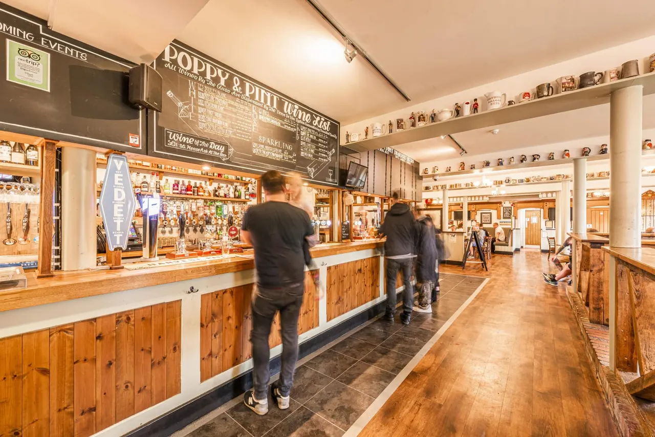 Visitors order drinks at a wooden-countered bar with shelves of bottles and merchandise displayed on walls.