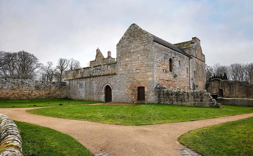 Medieval stone castle with crenellated walls set in grassy grounds with bare trees beyond.